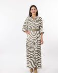 A woman modeling a long-sleeve button-front maxi shirt dress with a zebra print pattern in olive green and cream, featuring a matching belt with a gold buckle, set against a plain white studio background.
