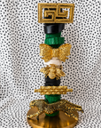 A tall decorative sculpture or fashion accessory stack featuring a gold Greek key plaque at the top, black and green wrapped sections, a gold bow tie, a gold bee, a gold patterned bar, and a gold leopard at the base, set against a black-and-white polka dot fabric backdrop.