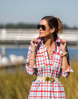 Woman in a red and white checkered dress standing in a grassy area with water and a dock in the background.