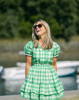 Woman in a green checkered dress standing by a body of water with boats in the background.
