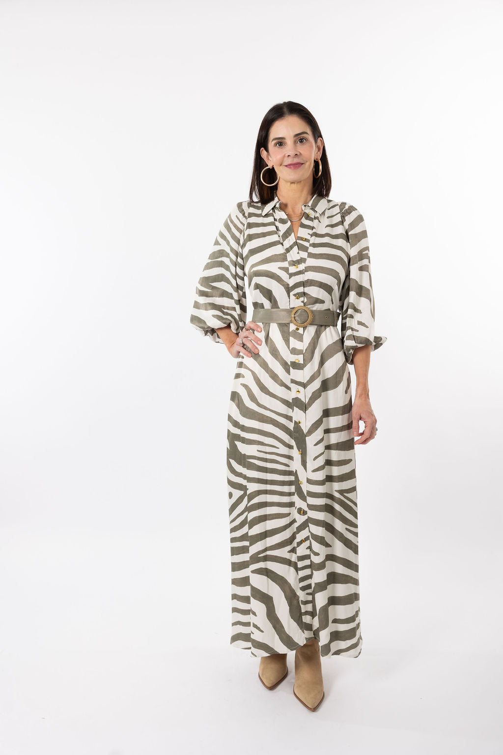 A woman modeling a long-sleeve button-front maxi shirt dress with a zebra print pattern in olive green and cream, featuring a matching belt with a gold buckle, set against a plain white studio background.