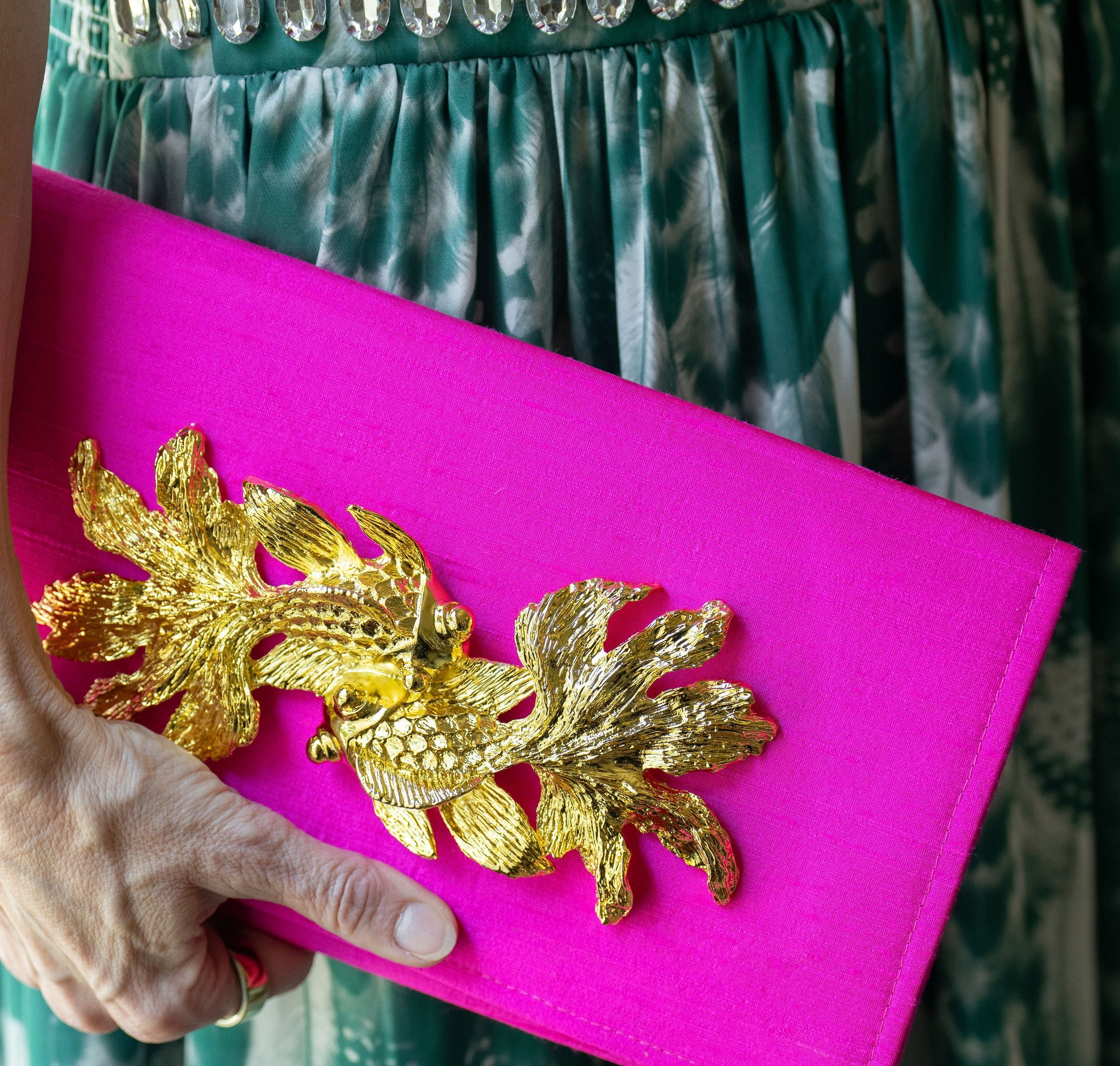 Pink clutch with gold leaf design held by a person wearing a green patterned dress.