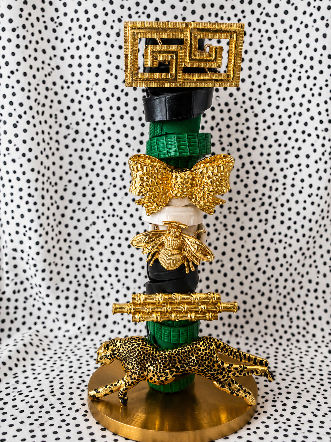 A tall decorative sculpture or fashion accessory stack featuring a gold Greek key plaque at the top, black and green wrapped sections, a gold bow tie, a gold bee, a gold patterned bar, and a gold leopard at the base, set against a black-and-white polka dot fabric backdrop.