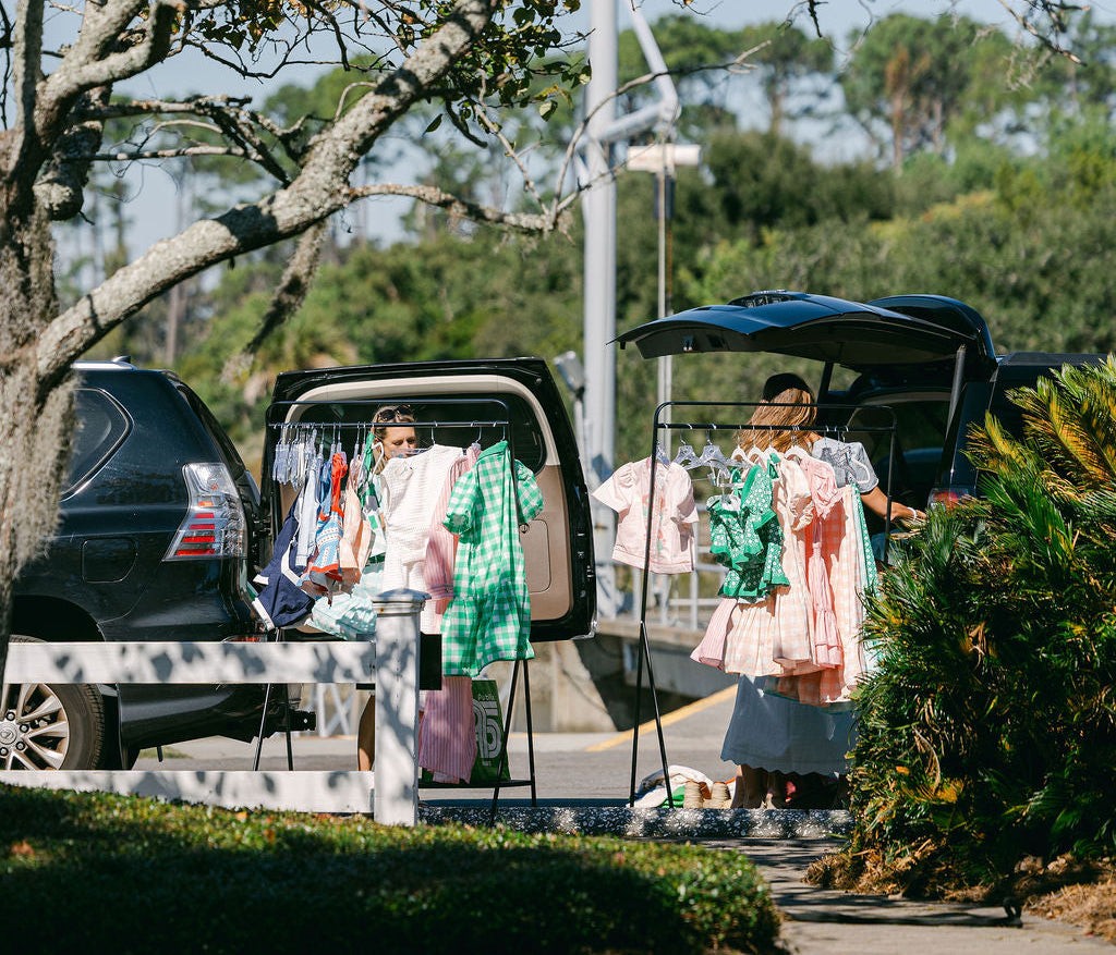 Two women standing next to open car trunks with clothing on a grassy area.