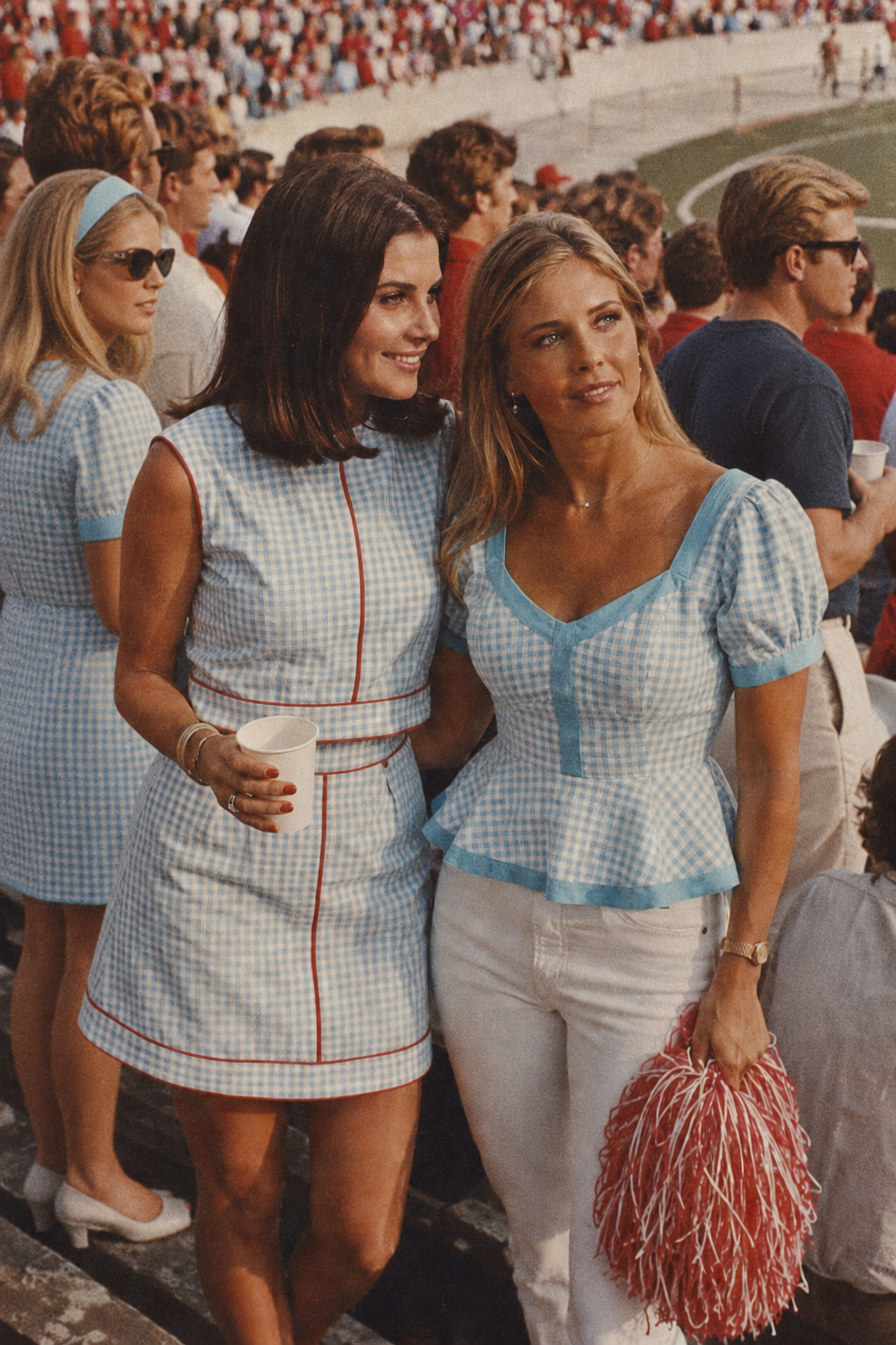 Two women standing in stadium seating at a sports event, wearing matching light blue and white gingham outfits. One woman wears a sleeveless mini dress with red piping, and the other wears a short-sleeve top with white pants and holds red-and-white pom-poms.