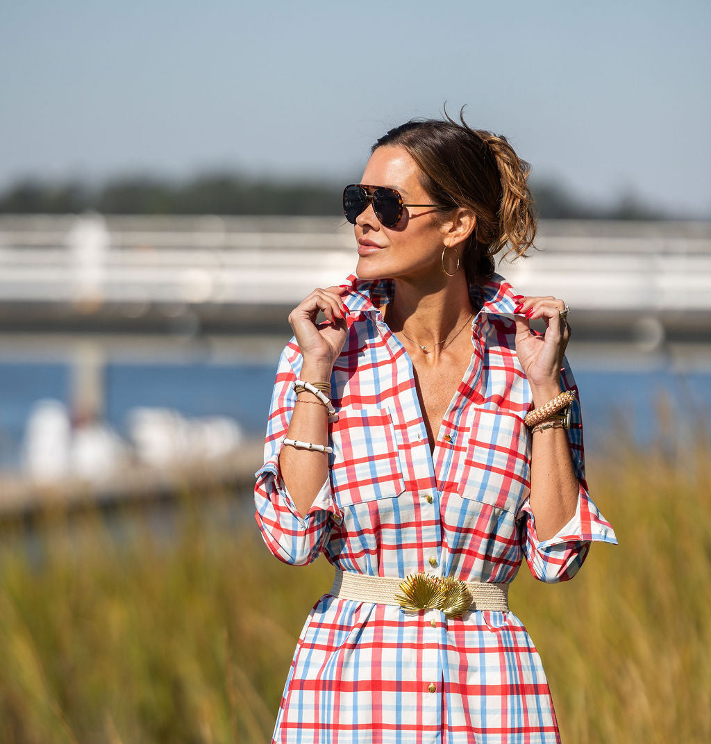 Woman in a red and white checkered dress standing in a grassy area with water and a dock in the background.
