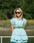 Woman wearing a light blue dress with green accents standing outdoors by a body of water.