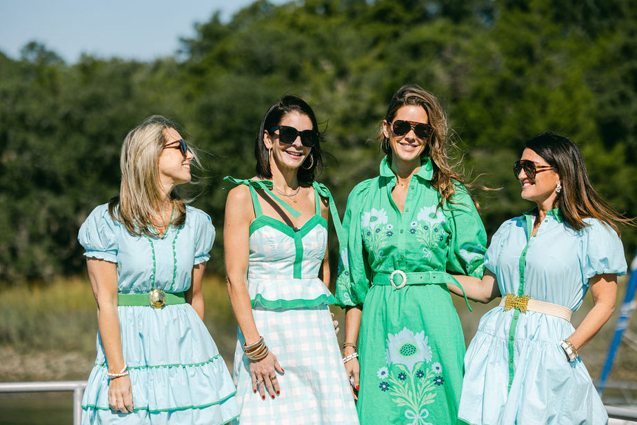 Four women in matching green and white dresses standing outdoors with trees in the background.