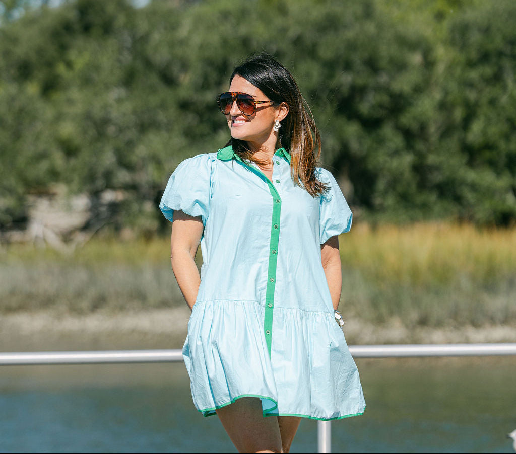 Woman in a light blue dress with green accents standing on a dock by a body of water.