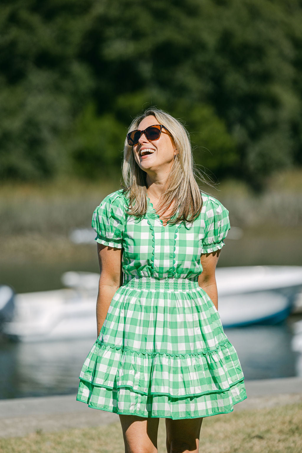 Woman in a green checkered dress standing by a body of water with boats in the background.