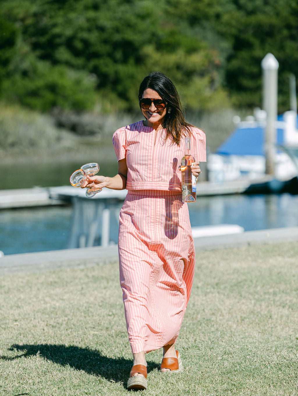 Woman in a pink outfit walking by a waterfront with trees and boats in the background